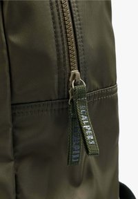 Mochila de nylon verde con una textura suave, que presenta una cremallera y un tirador de tela con letras azules. Diseño compacto para almacenamiento.