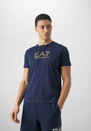 Camiseta de algodón azul marino con un logo dorado de "EA7 Emporio Armani". Mangas cortas, cuello redondo y una textura suave.