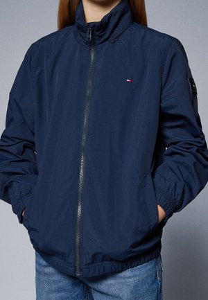 Person iført en navyblå jakke med lynlås og et lille rødt, hvidt og blåt logo på brystet samt hænderne i lommerne.