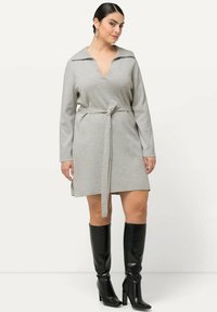 Abito in felpa grigio con scollatura a V profonda e maniche lunghe, legato in vita con una cintura coordinata, abbinato a stivali neri al ginocchio.