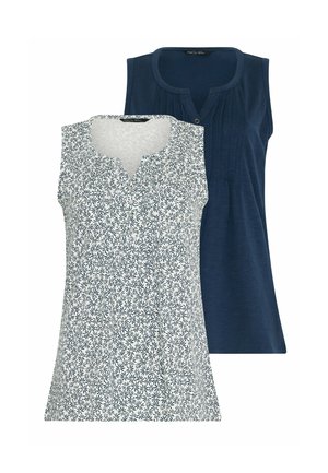 Frau mit lockigem Haar, die ein ärmelloses, gemustertes Oberteil, marineblaue Shorts und weiße Turnschuhe trägt, geht vorwärts vor einem schlichten Hintergrund.