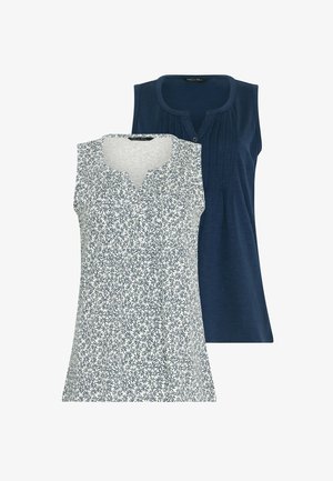 Vrouw met krullend haar, gekleed in een mouwloos topje met patroon, marineblauwe korte broek en witte sneakers, lopend naar voren tegen een effen achtergrond.