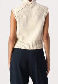 Top sin mangas de punto color crema con un cuello alto y un dobladillo acanalado; presenta un ajuste holgado y una tela texturizada. Combinado con pantalones oscuros.