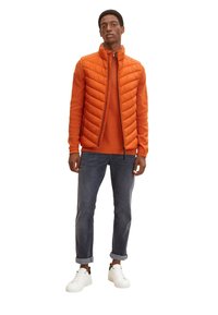 Doudoune orange vif avec un design matelassé, portée par-dessus un pull orange assorti. Assortie à un jean foncé retroussé et des baskets blanches.
