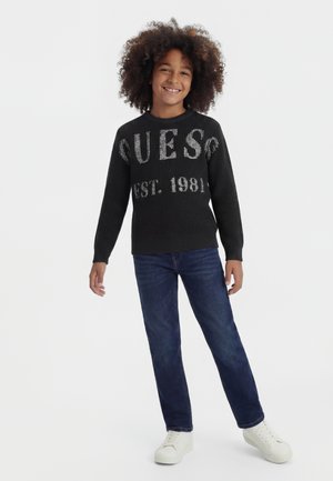 Osmijehnuto dijete s kovrčavom kosom, odjeveno u crni džemper "Guess Est. 1981", plave traperice i bijele tenisice, stoji ispred jednobojne pozadine.