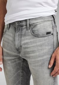 Jeans de mezclilla gris deslavado con un corte recto, diseño de cinco bolsillos, ligeras arrugas y una etiqueta negra de marca en el bolsillo delantero.