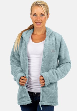 Frau mit blondem Haar, die eine hellblaue flauschige Reißverschlussjacke über einem weißen Shirt und dunklen Jeans trägt, steht vor einem schlichten Hintergrund.