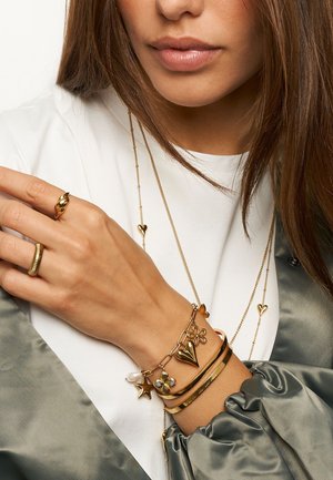 Femme portant des bagues en or, un bracelet de charme avec une étoile, une fleur, un cœur, une perle et deux colliers en chaîne dorés sur une chemise blanche et une veste en satin grise.