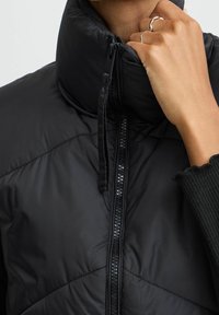Czarna kurtka puchowa z wysokim kołnierzem, przednim zamkiem, pikowanym wykończeniem i gładką powierzchnią. Szczegół ręki z srebrnymi pierścionkami na szyi.