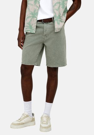 Férfi olíva zöld rövidnadrágban, fehér zokniban, bézs tornacipőben, fehér pólóban és nyitott gombolású ingben, pálmamintával.