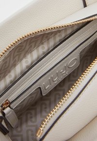 Intérieur d'un sac à main en cuir blanc montrant une poche zippée, des ferrures dorées et une doublure en tissu à motifs avec le nom de la marque visible.