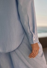 Camicia a righe azzurre e pantaloni con piccole paillettes, dotati di polsini con bottoni e orlo arrotondato. Tessuto morbido.