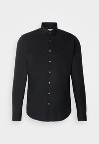 Chemise noire à manches longues avec col pointu, sept boutons visibles et logo discret sur la poitrine, sur fond blanc uni.