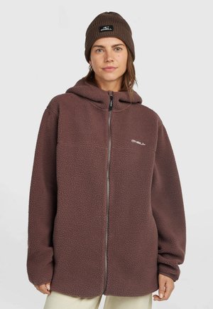 Vrouw die een bruine fleece met ritssluiting en bijpassende gebreide muts draagt, met een wit O'Neill-logo op beide artikelen, staand tegen een effen achtergrond.