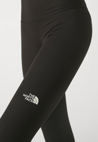 Czarne legginsy sportowe wykonane z rozciągliwego materiału, z białym logo na dolnej lewej nodze oraz wysokim pasem.