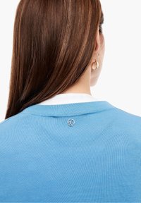 Modrý svetr s žebrovanou texturou a bílým límcem, s malým stříbrným logem blízko zadního výstřihu.