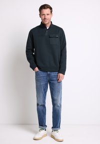 Uomo in piedi su uno sfondo bianco che indossa un pullover scuro con taschino, jeans blu arrotolati alle caviglie e sneakers bianche con accenti verdi.