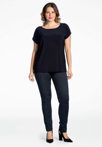Marinefarbene Kurzarmbluse mit lässiger Passform, kombiniert mit dunkelblauen Skinny Jeans und schwarzen High Heels. Einfaches Design, glatte Textur.