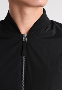 Černá bomber bunda s hladkým povrchem, žebrovaným límcem a stříbrným zipem. Malé logo viditelné na levé straně.