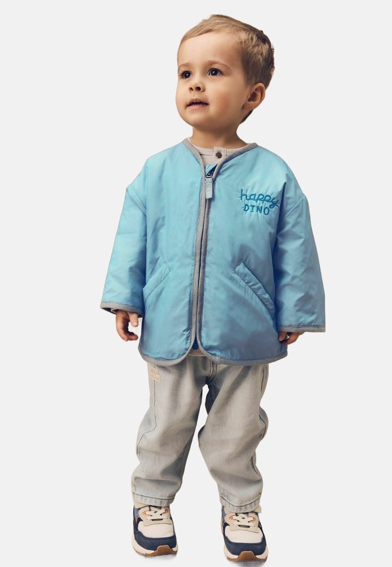 Ljusblå vadderad jacka med grå accenter, prydd med broderiet "happy DINO", matchad med ljusa denimbyxor och sportiga skor.