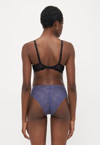 Zwarte kant bralette met verstelbare bandjes, gecombineerd met hoge paarse slips met een textuur golfpatroon, gezien vanaf de achterkant.