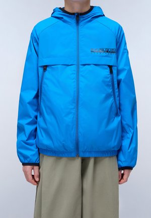 Personne portant une veste à capuche bleu vif zippée avec des fermetures éclair noires et des poignets élastiques, associée à un pantalon beige ample, sur fond uni.