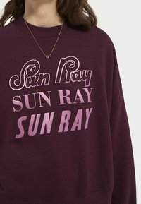 Sudadera de color burdeos con un gran texto rosa que dice "Sun Ray" en diferentes tipografías. Textura suave con cuello redondo y corte holgado.