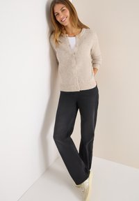 Beige neulecardigan, jossa on napit, yhdistetty mustiin suoriin housuihin ja kermaisiin lenkkareihin. Sujuva kangas, rento istuvuus ja rento muotoilu.