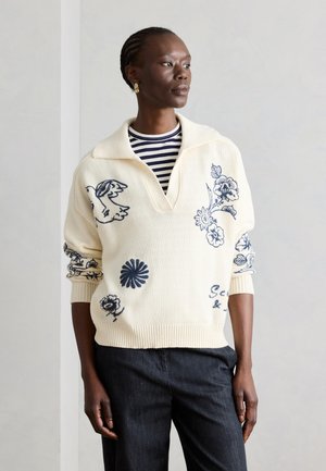 Femme aux cheveux tressés portant un pull crème avec broderies bleues de fleurs et d’oiseaux sur une chemise noire à rayures, et un pantalon foncé, sur fond uni.