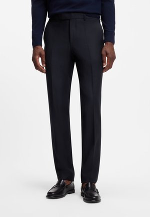 Homme portant un pantalon de costume noir sur mesure, des mocassins en cuir noir et un haut à manches longues sombre sur fond blanc.