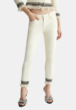 Pantalones vaqueros blancos ajustados con ribetes decorativos negros y dobladillo de flecos, hechos de denim, que cuentan con un bolsillo delantero y cierre con cremallera.