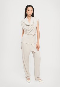 Ärmelloses Top mit Kapuzenkragen und locker sitzende Hose aus weichem beigen Stoff; spitze, cremefarbene Schuhe; minimalistische Gestaltung.