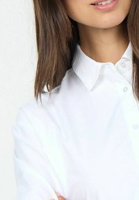 Camicia bianca con dettagli dei bottoni, realizzata in un tessuto liscio, mostra una parte della spalla e del petto di una persona con capelli scuri e dritti.