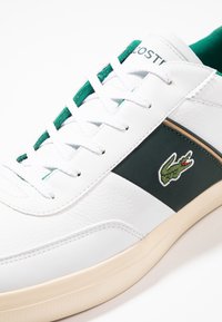 Bílé kožené tenisky se zelenými textilními detaily a tkaničkami. Na boku mají vyšívané logo v zelené a červené barvě a béžovou gumovou podrážku.
