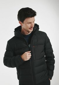 Czarna kurtka puchowa z zamkiem z przodu, kapturem i bocznymi kieszeniami. Posiada pikowaną strukturę oraz małe czerwone logo na kieszeni piersiowej.