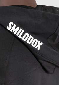 Černá mikina s kapucí vyrobená z měkkého materiálu má na pravém rameni bílé logo "SMILODOX". Jednoduchý design s kapucí a detaily šití.