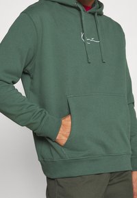 Grön hoodie i bomullsmix, med en framficka, ribbade mudd och nederkant, samt en vit logotyp på bröstet. Slät textur.