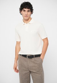 Νεαρός άνδρας με σκούρα σγουρά μαλλιά, φορώντας λευκό πουκάμισο polo και καφέ υφασμάτινο παντελόνι, στέκεται με το ένα χέρι στην τσέπη σε ουδέτερο φόντο.