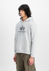 Lichtgrijze hoodie met een kangoeroezak, verstelbare trekkoord en een zwart logo op de borst. Gemaakt van zachte stof met ribgebreide manchetten.
