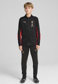Zwarte sportieve trainingspak met gouden accenten, met een ritsjaket met rode mouwen en logo, gecombineerd met zwarte tapered broek.