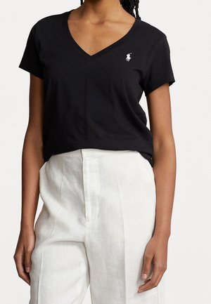 Kobieta ubrana w czarną koszulkę z krótkim rękawem i dekoltem w serek z białym logo oraz białe lniane spodnie z wysokim stanem, stojąca na jednolitym tle.