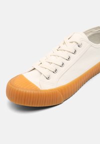 Canvas sneaker in off-white with contrast orange rubber sole. Features rounded toe, lace-up design, and metallic eyelets.