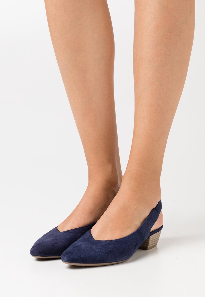 Femme portant des chaussures slingback en daim bleu marine à talon bloc bas, pieds nus, debout sur fond blanc.