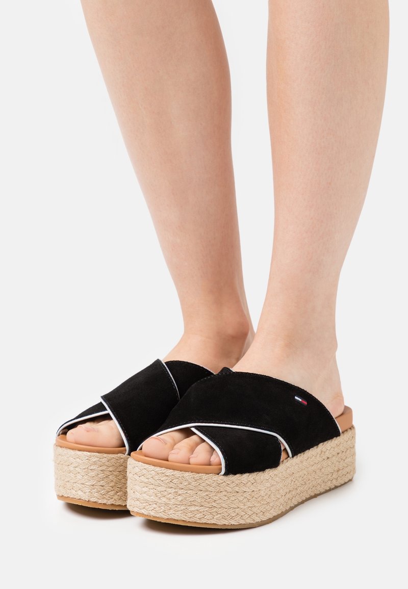 Pieds portant des sandales compensées en daim noir avec des brides croisées et des semelles en jute tissée sur fond blanc.