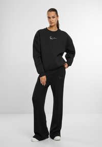 Felpa nera e pantaloni larghi in tessuto strutturato; presenta un piccolo logo bianco sul petto e sulla gamba del pantalone. Le scarpe sono bianche.