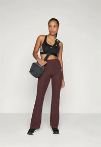 Czarny sportowy biustonosz z trzema białymi paskami, zestawiony z bordowymi spodniami o rozszerzanej nogawce i małą czarną torbą na ramię. Obuwie to czarne sneakersy.