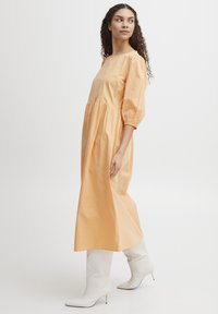 Perzikkleurige, enkellange jurk met puffmouwen en een gegatherde taille. Gepaard met witte, puntige hoge hakken.