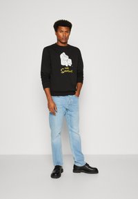 Sudadera negra de manga larga con un diseño gráfico de personajes de "Los Simpson", combinada con unos vaqueros azul claro y zapatos negros.