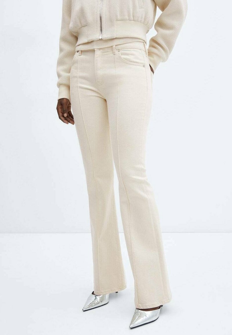 Pantalon beige évasé en denim doux, doté de poches avant et de coutures décoratives, assorti à des escarpins argentés brillants à bouts pointus.