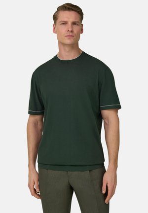 Homme portant une chemise à manches courtes vert foncé avec des bordures blanches sur les manches et le col, associée à un pantalon vert olive, debout devant un fond blanc.